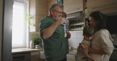 Baba emzik ile bebeği tutuyor, anne genç bir çocukla etkileşime geçiyor, aile bağlarını parlak, davetkar bir mutfak ortamında ön plana çıkarıyor.