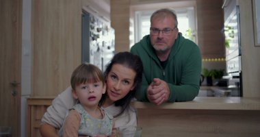 Anne ve büyük çocuk kameraya doğru eğilirken, baba mutfak tezgahından izliyor, sıcak ve iç içe geçen aile anı.