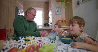 Çocuk kahvaltı yaparken, baba da bebeğe yemek hazırlarken, aile bağlarını ve sıcak mutfak ortamındaki yemek masasında bağlantıyı teşvik ediyor.