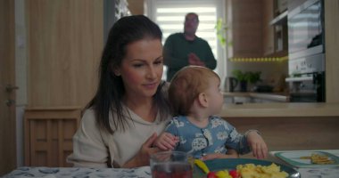 Anne kahvaltıda kucağında bebeği tutuyor, yeni yürümeye başlayan bir çocuk, mutfakta sıradan bir aile etkileşimi, sıcak ev ortamında günlük hayatın ayrıntıları.