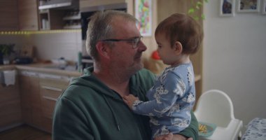 Baba kucağında bebekle, hassas bir anı paylaşıyor, rahat bir ev mutfağında göz teması kuruyor, anne arka planda yemek hazırlıyor.