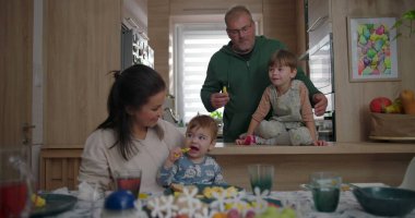 Anne ve bebek sıcak ve neşeli bir aile ortamında yemek masasında otururken baba ve büyük çocuk neşeyle etkileşime geçiyor.