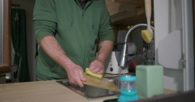 Eller mutfak lavabosunda musluk suyunun altında akan bir süngerle kesme tahtasını ovalayarak modern ev ortamında günlük temizlik işlerini tasvir ediyor.