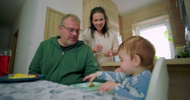 Anne ve baba yüksek sandalyede oturan bebekle iletişim kuruyor, baba bebeği besliyor, anne sıcak bir aile mutfağında gülümsüyor.