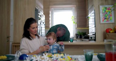 Anne bebeğe içkiyle yardımcı olurken baba mutfak tezgahına yaslanıyor, büyük çocuk sıcak aile ortamında sıcak bir yemek masasında babasıyla etkileşime geçiyor.