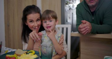 Anne, yemek masasında oturan büyük çocukla gülümserken baba mutfak tezgahına eğilir ve evde hassas bir aile etkileşimi yakalar.