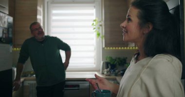 Kadın elinde bardak tutarken sıcak bir şekilde gülümsüyor. Mutfakta duran bir adamla sohbet ediyor. Dostça ve rahat bir atmosferi teşvik ediyor.