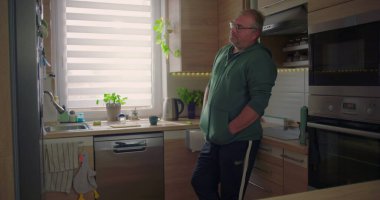 Adam mutfak tezgahına yaslanıyor, elleri cepte, modern mutfak arka planı, günlük rutin işler, düşünceli anlar, yeşillik ve dekorasyon.