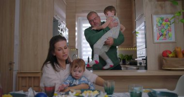 Baba, daha yeni yürümeye başlamış bir çocuk, annesiyle yemek masasında oturup, bir aile mutfağında neşe ve bağ kurarken neşe içinde büyük bir çocuğu kaldırıyor.