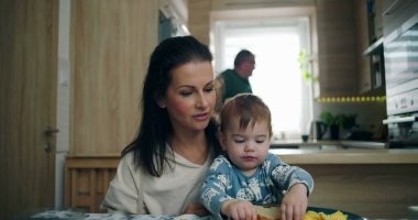 Anne, yemek masasında bebeğini besliyor. Bebek, sıcak ve keyifli bir aile ortamını paylaşırken yemeği keşfediyor.