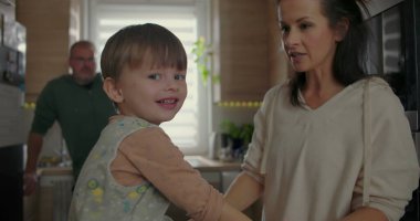 Anne küçük bir çocuğu mutfaktan alıyor, sevgi ve ilgi gösteriyor, arka planda bir adam tezgaha yaslanıyor, sıcak aile anı içeride.