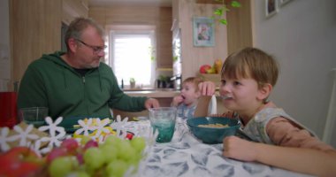 Çocuk kahvaltı yaparken, baba da bebeğe yemek hazırlarken, aile bağlarını ve sıcak mutfak ortamındaki yemek masasında bağlantıyı teşvik ediyor.