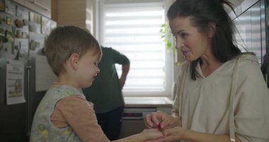 Anne elinde küçük bir tatlı tutuyor, mutfakta çocuğuna gösteriyor, arka planda gelişigüzel eğilen bir adam, öğrenme ve merak anı.