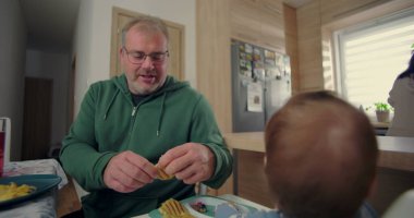 Babam kahvaltıda yürümeye başlayan çocuğa yemek hazırlıyor, ilgiye ve ilgiye odaklanıyor, sıcak ev mutfağında samimi bir aile ortamı yaratıyor.