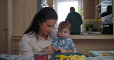 Anne yemek masasında bebeği besler, evde sıcak ve samimi bir aile ortamında renkli yiyeceklerle ilgilenir.