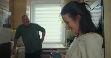 Mavi kupa tutan bir kadın, modern bir mutfakta sıradan bir adamla konuşuyor, sıcak bir sohbete girişiyor, ev ortamında rahat ve arkadaşça bir etkileşim içinde.