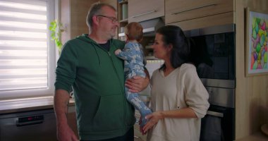 Anne ve baba kucağında bebekle gülümsüyor, modern mutfakta sıcak ev havalarıyla aile anlarının tadını çıkarıyorlar.