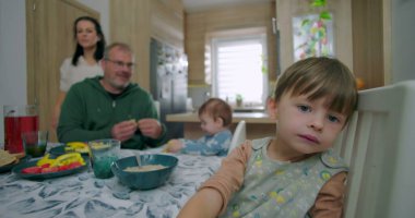 Genç bir çocuk yemek sandalyesine yaslanmış meraklı görünüyor, babası küçük çocuğu yüksek sandalyede besliyor, annesi arka planda ayakta gülümsüyor, aile mutfakta kahvaltı için toplanıyor..