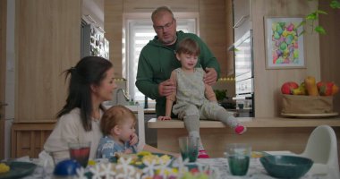 Anne, mutfak tezgahında oturan büyük çocuğa yemek sunarken baba yakınlarda duruyor. Bebek, annesinin kucağında yemek masasındaki aile bağlarını inceliyor..