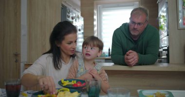 Anne ve büyük çocuk yemek masasında bir anı paylaşırken, baba mutfak tezgahından gözlemler, bir aile ortamında bağ ve sıcaklığı teşvik eder.