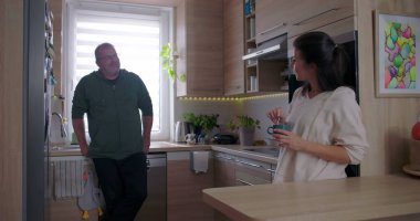Mutfak tezgahına dayanmış bir adam elinde kahve fincanı tutan bir kadınla konuşuyor. İyi aydınlatılmış, ahşap iç mekânlı, modern bir mutfakta sıradan bir sohbet.