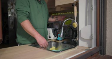 Modern bir mutfak içinde bulaşık yıkayan bir adam, lavabonun yanında elleriyle bir kase temizliyor, rahat bir ev ortamında günlük ev işleri yapıyor.
