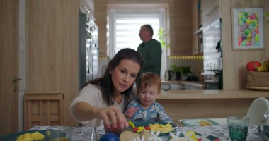 Anne ve bebek kahvaltı masasında, neşeli bebekle beslenme anını paylaşıyorlar, aile mutfakta basit anların tadını çıkarıyor, paylaşılan yemek sırasında gelişigüzel bağ kuruyorlar.