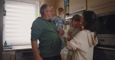 Modern bir mutfakta bir erkek bebeğini tutarken bir anne büyük bir çocukla etkileşime geçip sıcak ve samimi aile dinamikleri yakalar.