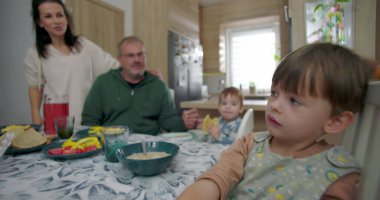 Çocuk, hayır der gibi başını sallıyor, muhtemelen yemeği reddediyor ya da kendini soyutlanmış hissederken, babası bebek bebekle yüksek sandalyede besleniyor, aile mutfakta toplanıyor, anne de arka planda..