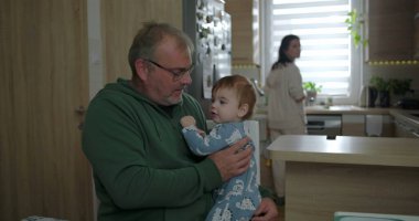 Baba kucağında bebek, anne mutfakta çalışırken hassas bir anı paylaşıyor sıcak ve sevgi dolu bir ev ortamında aile içi etkileşimi.