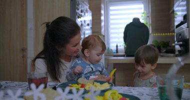 Anne kahvaltıda bebeğe biber uzatıyor, sıcak mutfak ortamında aile olarak yiyeceği ve kaynaşmayı paylaşıyor ve etkileşimi besliyor.