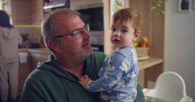 Baba, yeni yürümeye başlayan çocuğu mutfakta tutuyor, aile içinde samimi bir an, kucağında bebek varken meraklı görünüyor, arka plan yemek ve mutfak unsurlarını gösteriyor, sıcak aile ortamını gösteriyor.