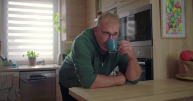 Modern mutfakta içki içen bir adam, rahat ev anı, günlük sabah rutini, ev ortamında yeşillik ve güneş ışığı, rahat yaşam tarzı.