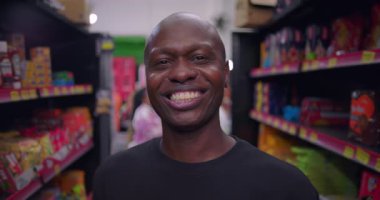 Market koridorunda gülümseyen Afrikalı bir adam, kameraya yakın, renkli ürün raflarıyla çevrili, siyah tişörtlü ve doğrudan kameraya bakıyor.