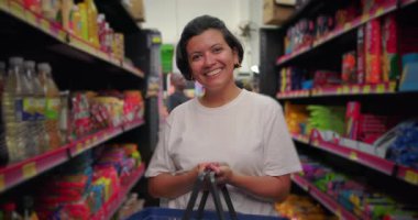 Market koridorunda alışveriş sepetiyle gülümseyen Latin kadın, doğrudan kameraya bakıyor, renkli paketlenmiş yiyecekler ve ev ürünleriyle çevrili.