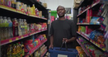 Market koridorunda elinde alışveriş sepetiyle dikilen Afrikalı adam, sakin bir ifadeyle kameraya bakıyor, her iki tarafta yiyecek rafları ve ev eşyaları var.