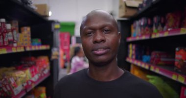 Market koridorunda sakin bir ifadeye sahip Afrikalı adam renkli ürünler ve arka plandaki raflarda paketlenmiş yiyeceklerle kameraya bakıyor.