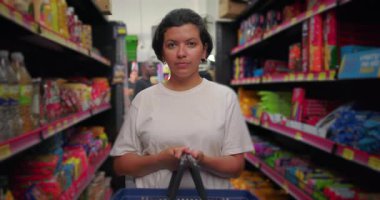 Süpermarkette elinde alışveriş sepetiyle dikilen Latin bir kadın. Ciddi bir ifadeyle kameraya bakıyor. Etrafı renkli market eşyalarıyla çevrili.