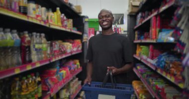 Market koridorunda elinde sepet tutarken gülümseyen Afrikalı bir adam renkli ürün raflarının önünde duruyor, arka planda gezinen bir kadın.