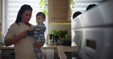 Anne mutfakta gülümserken bebeği kucağına alıyor, günlük ebeveynlik rutini sırasında çocukla meşgul oluyor, ev ortamında sıcak ve sıradan bir aile sahnesi.