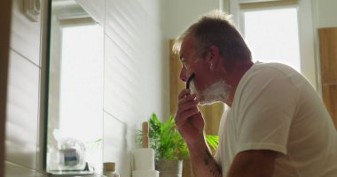 Ustura ve köpükle tıraş olan, banyoda duran, favorinin olduğu yere odaklanan, çerçevede ayna yansıması, kişisel bakım, sakin ve düşünceli bir ifade.