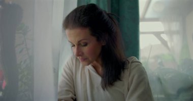 Bir kadın pencereden dikkatlice dışarı bakıyor, çenesini elinin üzerine koyuyor, derin bir tefekkür içinde, ev içi detaylar ve yeşil perdelerle çevrili.