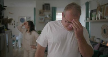 Tartışma sırasında elleri hüsran içinde yüzünü kapatan erkek, oturma odasında arka planda kalan kadın, dramatik ilişki çatışması, duygusal mücadeleye odaklan.