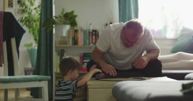 Baba ve çocuk sehpanın üzerinde oyuncak trenle oynuyorlar, anne kanepeye oturuyor, küçük beyaz bir köpek tutuyor, rahat bir aile anı, bağ kurma ve ev ortamında oyun zamanı.