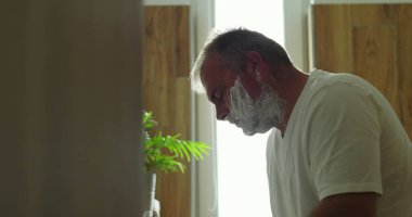 Tıraş makinesiyle yüzünü traş eden, bakım rutininde hassasiyet ve özgüvene odaklanan, modern ve temiz banyo ortamının penceresinden yansıyan parlak ışık.