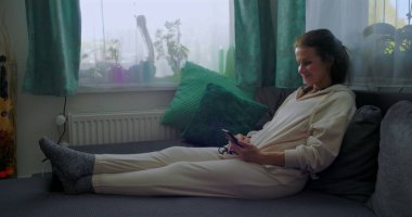 Kanepede uzanan, akıllı telefon kullanan, rahat oturma odasında rahatlayan, yumuşak yastıklar ve doğal ışık ile çevrili, kapalı alanda sakin bir aktivite.