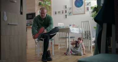 Babam botlarını bağlarken küçük bir çocuk elinde oyuncak bir arabayla yerde oturuyor, aile yemek odasında ahşap döşemeler ve dekoratif unsurlarla.