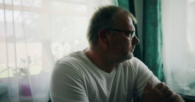 Pencere kenarında oturan bir adam, kollarını kavuşturmuş, yan tarafa derin derin bakıyor, sakin ve sakin bir an, sakin bir ortamda perdelerle çerçevelenmiş