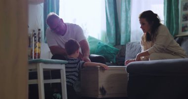 Baba masada genç bir çocukla oynarken anne gülümsüyor, aile içinde sıcak bir oturma odasında yakınlık kuruyor ve aktivitelerini paylaşıyor.