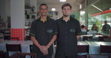 İki restoran garsonu yemek salonunda beklerken gülümsüyor. Elleri katlı ve arkada. Cam duvar ve arka planda görünen masalar.